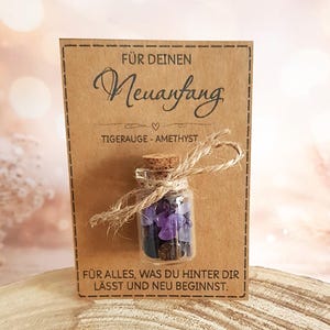 Könnte beinhalten: Eine kleine Glasflasche gefüllt mit lila und braunen Edelsteinen, mit Bindfaden gebunden und einem Korken verschlossen. Die Flasche ist an einer braunen Karte mit deutschem Text befestigt. Auf der Karte steht "FÜR DEINEN Neuanfang" und "TIGERAUGE - AMETHYST".