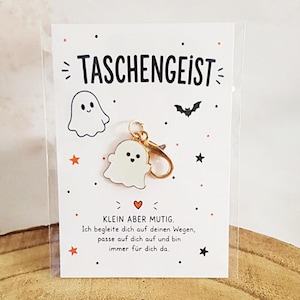 Könnte beinhalten: Ein weißer, geisterförmiger Emaille-Anhänger mit goldenem Karabinerverschluss auf einer weißen Präsentationskarte. Die Karte zeigt den Text TASCHENGEIST, eine Geisterillustration, eine Fledermaus, Sterne und den Text KLEIN ABER MUTIG.