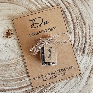 Könnte beinhalten: Eine kleine Glasflasche mit Korken, mit Bindfaden gebunden, und einem kleinen Etikett mit der Aufschrift "Tigerauge". Die Flasche befindet sich auf einer braunen Karte mit den Worten "Du SCHAFFST DAS!" und "WEIL DU MEHR STÄRKE HAST, ALS DU GLAUBST."
