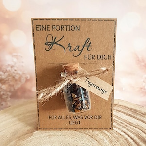 Könnte beinhalten: Ein kleines Glasfläschchen gefüllt mit Tigeraugensteinen, mit Bindfaden an einer Kraftpapierkarte befestigt. Die Karte trägt den Text "EINE PORTION Kraft FÜR DICH" und "FÜR ALLES, WAS VOR DIR LIEGT."