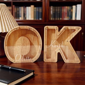 May include: Two wooden letter-shaped money banks, an "O" and a "K". The "O" has the name "Odette" in script, and the "K" has "Keith". The banks are made of light-colored wood and have a clear front panel. A pen and notebook are in the foreground.