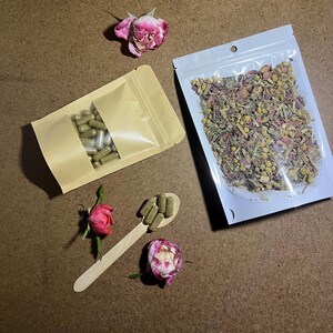 以下が含まれることがあります： ハーブサプリメントと乾燥ハーブの俯瞰図。カプセルは茶色の紙袋に入っており、乾燥ハーブは透明なビニール袋に入っています。木のスプーンにはカプセルが入っており、ピンクのバラが散らばっています。