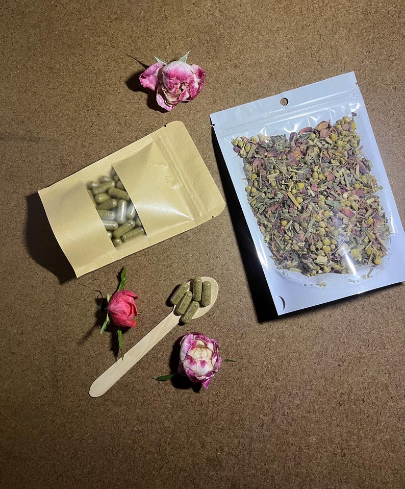 以下が含まれることがあります： ハーブサプリメントと乾燥ハーブの俯瞰図。カプセルは茶色の紙袋に入っており、乾燥ハーブは透明なビニール袋に入っています。木のスプーンにはカプセルが入っており、ピンクのバラが散らばっています。