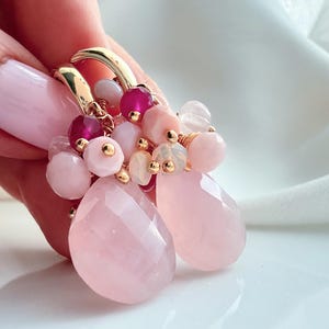 Op de afbeelding: Een paar gouden oorringen versierd met clusters van gefacetteerde lichtroze druppelvormige stenen, kleine ronde roze kralen en diep magenta kralen. De oorbellen zijn voorzien van delicaat gouddraad en een mix van opake en doorschijnende edelsteentexturen.