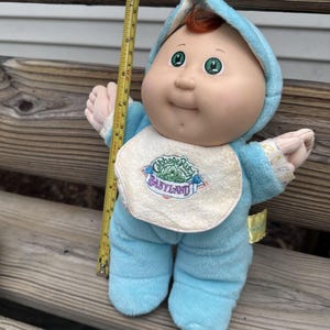 May include: A Cabbage Patch Kids Babyland doll with red hair, green eyes, and a light blue hooded onesie. A cream-colored bib features the embroidered text Cabbage Patch Babyland. A yellow measuring tape shows the doll is approximately 10 inches tall.