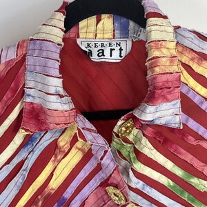 May include: A red jacket featuring a collar and front panels decorated with vertical, frayed-edge fabric strips in yellow, purple, and blue tie-dye patterns. The garment includes gold-toned buttons and a label reading Keren Hart.