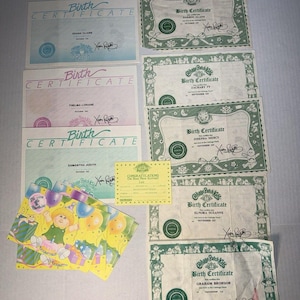 May include: A collection of Cabbage Patch Kids birth certificates and birthday cards. The documents feature green or blue borders, printed text, and signatures. Several cards display a cartoon child with balloons and a cake, alongside a small yellow card.