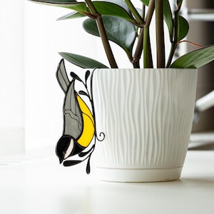 May include: A white, textured ceramic pot with a live plant. The plant has dark green leaves and reddish stems. A decorative stained glass bird with yellow, gray, and black accents is attached to the pot.