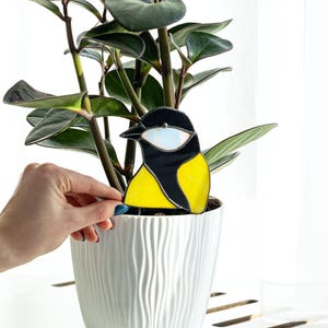 May include: A stained glass bird decoration in a white pot with a green plant. The bird is yellow, black, and white. The pot is white with a textured surface. The plant has dark green leaves.