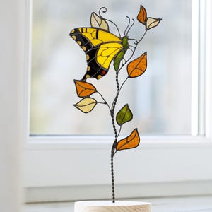 May include: A stained glass butterfly sculpture with yellow, black, and red details, on a metal stem with colourful leaves. The sculpture is mounted on a small, round wooden base, ideal for home decor.