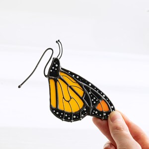 May include: A stained glass monarch butterfly wing with black outlines, white dots, and a vibrant orange and yellow pattern. The wing is held by a hand against a white background. The butterfly wing has a wire for hanging.
