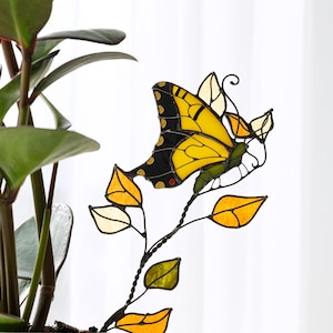 May include: A stained glass butterfly decoration with yellow and black wings, perched on a metal stem with yellow and green leaves. The butterfly is set against a white background, near a potted plant.