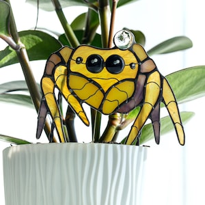 Op de afbeelding: Een gebrandschilderde krabbenplantenstaak in een witte pot met een groene plant. De krab is geel met zwarte ogen en bruine en paarse accenten. Bovenop de kop van de krab zit een heldere glazen bol.