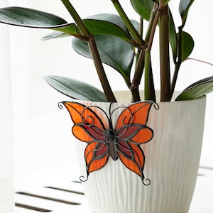 May include: A white ceramic planter with a textured surface, holding a green plant with dark stems. An orange and red stained glass butterfly decoration is attached to the planter. The butterfly has black metal detailing.
