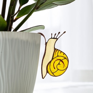 May include: A decorative stained glass snail plant stake. The snail has a yellow body with a brown outline and a spiral shell. The stake is attached to a white ceramic pot with a ZZ plant.
