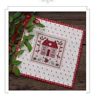 May include: A cross-stitch mini quilt with a cottage design, red roof, and heart details. The quilt has a white background with red and white patterned borders, decorated with green leaves and red ribbon. The text reads "The Cottage on the Corner".
