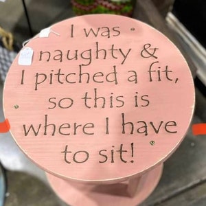 May include: A small, round, pink wooden stool with the text "I was naughty & I pitched a fit, so this is where I have to sit!" in dark brown lettering. The stool has a circular top and a pedestal base.