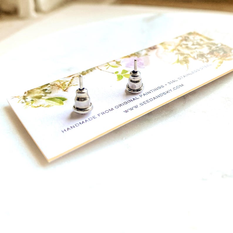 May include: Two silver-colored stud earrings with a tiered design are displayed on a white card with floral accents. The card reads "HANDMADE FROM ORIGINAL PAINTINGS - 316L STAINLESS STEEL" and "WWW.SEEDANDSKY.COM".