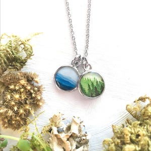 May include: A silver necklace with two round pendants. The pendants feature watercolor paintings of a mountain range and a forest.
