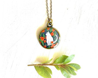 May include: A white cat sitting in a field of red poppies, surrounded by green leaves. The image is on a round, bronze pendant with a chain.