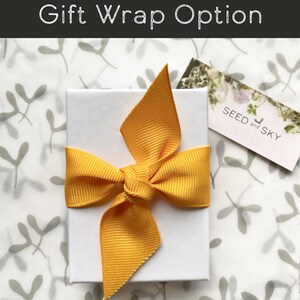 May include: A white gift box tied with a bright yellow ribbon. The box is on a patterned background with the text "Gift Wrap Option" at the top. A small tag with the text "SEED and SKY" is visible.