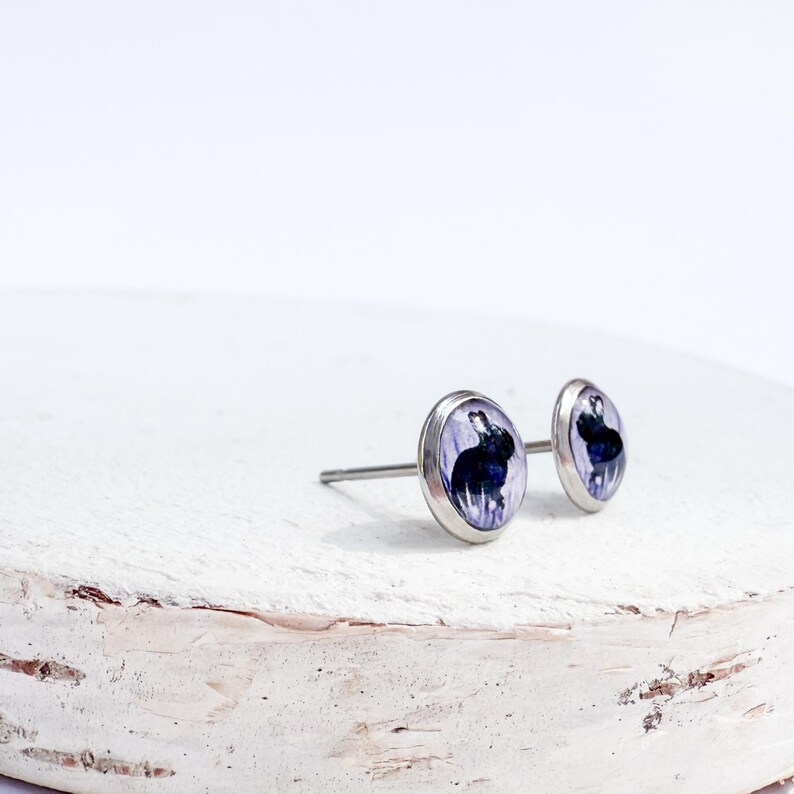 May include: A pair of silver stud earrings. Each earring features a small, oval-shaped design with a dark blue and purple abstract image. The earrings are displayed on a white, textured surface.