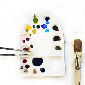 May include: A white, kidney-shaped artist's palette filled with various colors of paint, including yellow, blue, green, and brown. Two thin paint brushes and one large brush are also present, suggesting a painting setup. The brushes have wooden handles and metal ferrules.