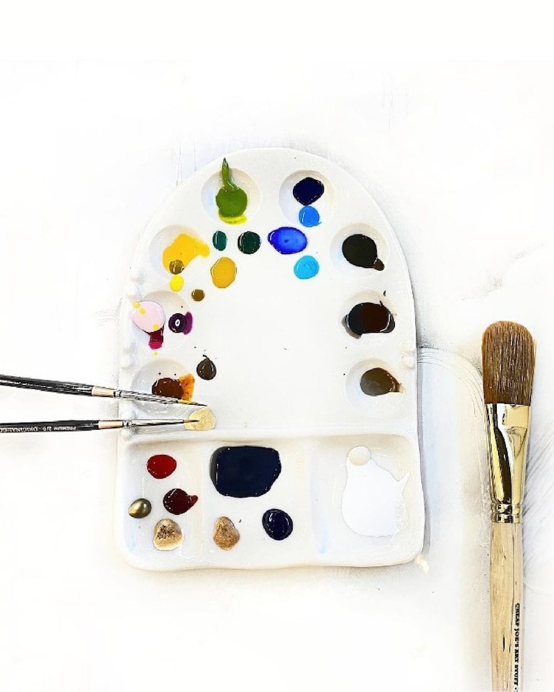 May include: A white, kidney-shaped artist's palette filled with various colors of paint, including yellow, blue, green, and brown. Two thin paint brushes and one large brush are also present, suggesting a painting setup. The brushes have wooden handles and metal ferrules.