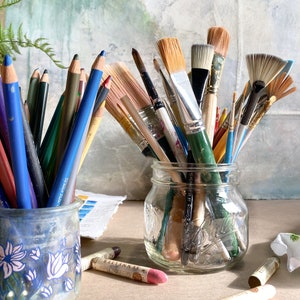 May include: A collection of art supplies, including colored pencils in a blue floral ceramic jar and various paintbrushes in a clear glass jar. Pastel chalks are scattered on the surface. The background features a blurred painting.