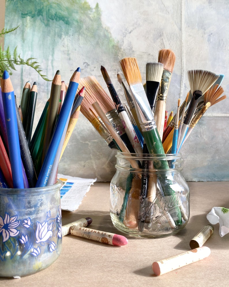 May include: A collection of art supplies, including colored pencils in a blue floral ceramic jar and various paintbrushes in a clear glass jar. Pastel chalks are scattered on the surface. The background features a blurred painting.
