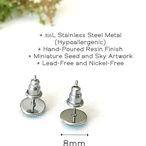 May include: Two stainless steel stud earrings with a hand-poured resin finish. The earrings are 8mm (roughly 1/3") in diameter and feature miniature seed and sky artwork. The text indicates they are lead-free and nickel-free.