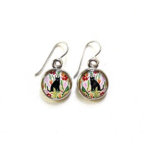 May include: A pair of silver-toned dangle earrings. Each earring features a round, floral design with a black cat illustration. The background includes red, pink, and orange flowers with green foliage, set against a white backdrop.
