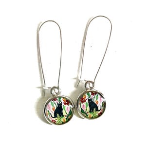 May include: A pair of silver-tone drop earrings. Each earring features a round charm with a black cat sitting among colorful flowers. The flowers are red, pink, and green against a white background. The earrings have long, curved ear wires.
