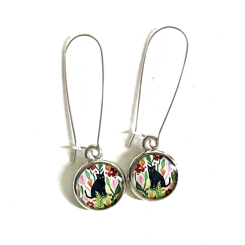 May include: A pair of silver-tone drop earrings. Each earring features a round charm with a black cat sitting among colorful flowers. The flowers are red, pink, and green against a white background. The earrings have long, curved ear wires.