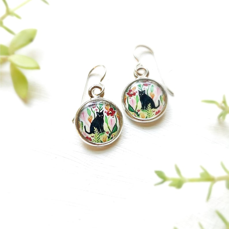 May include: A pair of silver-toned dangle earrings. Each earring features a round, glass-covered image of a black cat sitting in a colorful garden of flowers and foliage. The earrings have a simple hook design.
