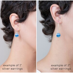 May include: Silver earrings with a round, blue design. The image shows two examples: one with 1-inch earrings and another with 2-inch earrings. The earrings have a hook closure and a bright blue, glittery interior.