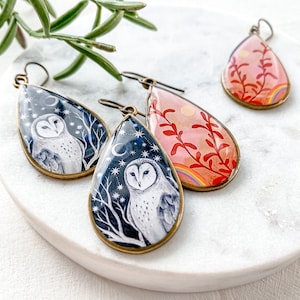 Könnte beinhalten: Tropfenförmige Ohrringe mit einzigartigen Designs. Zwei Ohrringe zeigen eine weiße Eule vor einem dunkelblauen Sternenhimmel, während ein anderer einen rosa Hintergrund mit einem Regenbogen und roten Pflanzenillustrationen hat. Die Ohrringe haben bronzefarbene Metallrahmen.