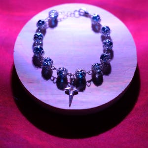 May include: A silver bracelet with blue gemstone beads and a star pendant. The jewellery is displayed on a wooden disc against a red background. The bracelet is made of linked metal and features multiple round blue beads.