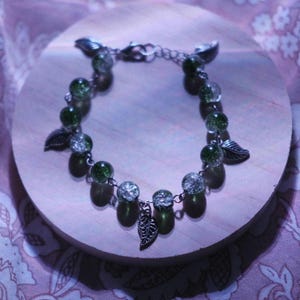 May include: A silver bracelet with green and clear glass beads, and silver leaf charms. The bracelet is displayed on a light wooden surface, against a patterned purple background. The bracelet has a lobster clasp closure.