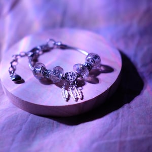 May include: A silver bracelet with clear, faceted beads and a dangling dreamcatcher charm. The bracelet is displayed on a round, light-coloured wooden surface, set against a soft, purple-toned background.