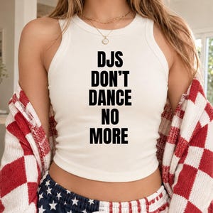 May include: A white ribbed tank top featuring the black text DJS DON'T DANCE NO MORE printed on the front. The garment is worn with a red and white checkered cardigan and blue shorts with a white star pattern, paired with a gold chain necklace.
