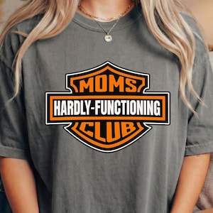 May include: Grey t-shirt featuring the text "MOMS HARDLY-FUNCTIONING CLUB" in black, orange, and white. The shirt has a relaxed fit and short sleeves. A woman is wearing the shirt and holding a mug.