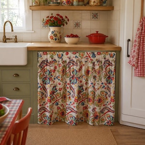 Peut inclure: Un rideau de placard de cuisine arborant un motif floral et cachemire aux tons rouge, bleu, vert et jaune sur fond crème. Le tissu est froncé sur le haut et recouvre la façade d'un meuble de cuisine en bois de couleur verte.