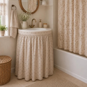 Op de afbeelding: Een badkamermeubel met een beige stoffen rok met een klein donker bloemenpatroon, een witte wastafel, een messing kraan, een ronde houten spiegel en een bijpassend beige bloemrijk douchegordijn aan een stang boven een wit bad.
