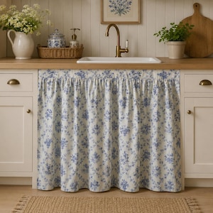 Könnte beinhalten: Ein weißer Spülenvorhang aus Stoff mit blauem Blumenmuster im Toile-de-Jouy-Stil und gerafftem Oberrand. Der Vorhang ist unter der Spüle angebracht und verdeckt die Schranköffnung mit einem plissierten Design und wiederkehrendem Blumenmotiv.
