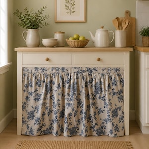 Op de afbeelding: Een witte houten keukentafel met twee lades en een geplooid stoffen gordijn met een blauw bloemenpatroon. Op het bovenblad staan een keramische kan, kommen, een pot met deksel, een rieten mand met groene appels en een theepot.