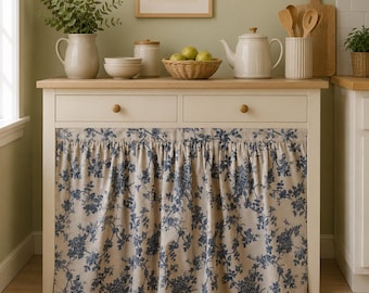 May include: A white wooden kitchen console table featuring two drawers and a gathered fabric curtain with a blue floral pattern. The top surface holds a ceramic pitcher, bowls, a lidded jar, a wicker basket of green apples, and a teapot.