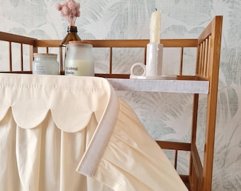 May include: A cream-colored fabric skirt with a scalloped top edge and a hook-and-loop fastener strip, attached to a wooden changing table. The table holds a glass jar with a white label, a brown glass bottle with dried flowers, and a candle.