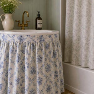 Könnte beinhalten: Ein Waschbeckenunterschrank-Vorhang mit blau-weißem Blumenmuster. Das Waschbecken hat ein weißes Becken, eine Messingarmatur und einen braunen Glasseifenspender. Ein weißer Krug mit weißen Blumen steht auf der Ablagefläche des Waschbeckens.