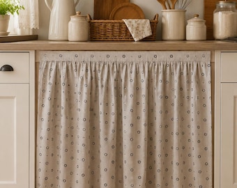 May include: A beige kitchen sink skirt featuring a repeating pattern of small dark brown flowers. The fabric is gathered at the top with a straight header, covering the lower cabinet area beneath a wooden countertop in a kitchen setting.